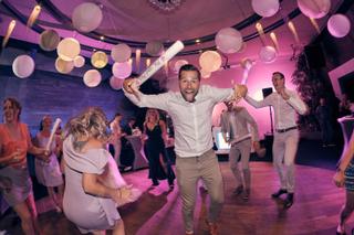Bruidegom Michiel in witte overhemd springt joelend op de dansvloer onder roze licht en papieren bollen, bruid Angelique en g