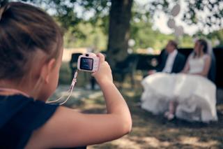 Kleine bruidsjonker fotografeert het bruidspaar met roze kindercamera onder de bloeiende bomen - Trouwshoot Stefan Segers
