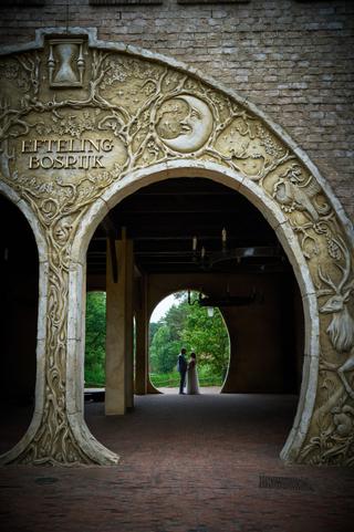 Annemarie en Jan onder de ornamentale boog in Bosrijk, Efteling. Het bruidspaar poseeert elegant in het groene landschap - Tr