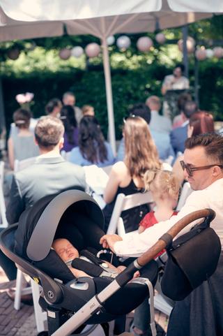 Pasgeboren baby in kinderwagen tijdens bruiloft op terras van De Schaapskooi in Delft, familie en gasten op achtergrond - Tro