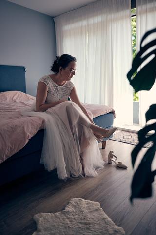 Bruid Evelien in witte trouwjurk zit op bed in slaapkamer, moment voor ceremonie, trouwreportage De Schaapskooi Delft - Trouw