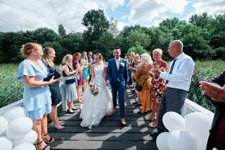 Bruidspaar Tessa en Jeroen wandelt onder applaas van gasten in groen landschap, trouwreportage Het Rieten Dak Delft - Trouwsh