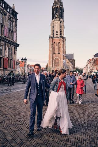 Bruidspaar Harm-Jan en Huszár loopt hand in hand over Delftse markt met Nieuwe Kerk op achtergrond, trouwreportage Delft - Tr
