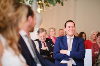 Bruidegom David kijkt glimlachend naar bruid Sandra tijdens ceremonie, gasten in zaal Het Spaansche Hof Den Haag - Trouwshoot