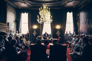 Trouwceremonie in prachtig stadhuis Den Haag, bruidspaar voor gouden kroonluchter, getuigen en gasten in elegant interieur -