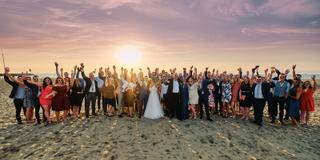 Bruidspaar Stefan en Victoria met alle gasten armen omhoog op strand Waterreus, zonsondergang trouwreportage - Trouwshoot Ste
