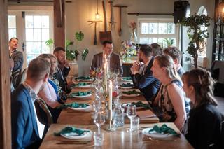 Ilse en Robbert aan de feesttafel met gasten bij Boerderij Burgersdijk in De Lier, warm licht en groene decoratie - Trouwshoo