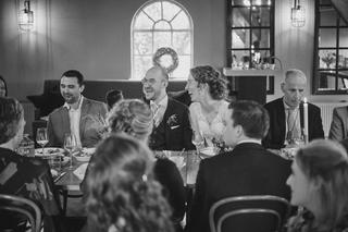 Ilse en Robbert lachen samen aan tafel tijdens hun bruiloftsfeest op Boerderij Burgersdijk, omringd door gelukkige gasten - T