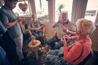 Bruidspaar Silvia en Patrick delen een lach met gasten op Beachhouse Kijkduin, hartelijk moment in stralend daglicht - Trouws
