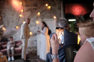 Gast fotografeert bruidspaar met smartphone tijdens trouwreportage, gloeiende lichtkransen op historische locatie - Trouwshoo