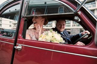Bruidspaar Maddy en Wijnand wuiven blij uit rode vintage auto, bruiloft Villa Ockenburg Naaldwijk, trouwreportage - Trouwshoo