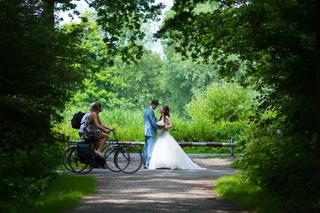 Bruidspaar Richard en Danielle kussen op fietspad onder groen bladerdak, vrouw op fiets passeert - Trouwshoot Stefan Segers