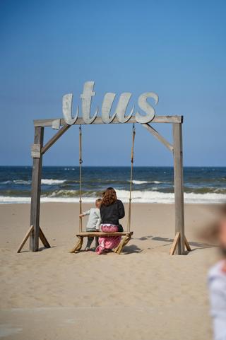 Elise en Erik op de schommel bij Beachclub Titus in Kijkduin met het glinsterende strand en zee op de achtergrond - Trouwshoo