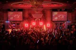 Bruidspaar en gasten dansen op feestdans onder rode neonverlichting, luxe zaal met kristallen kroonluchter, trouwfeest met DJ
