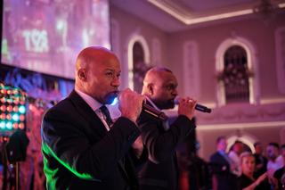 Twee mannen zingen in microfoon op trouwfeest, paars verlichte feestzaal met dansende gasten, festive bruiloftsfeest - Trouws