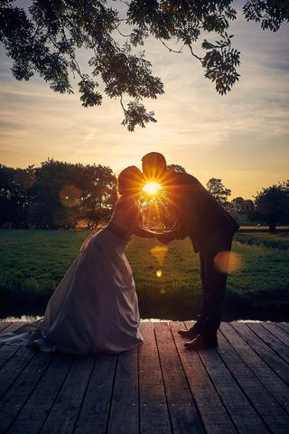 Bruidspaar kust bij zonsondergang met zon tussen hoofden, romantische trouwfoto op houten steiger - Trouwshoot Stefan Segers