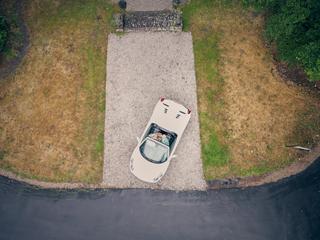 Bruidspaar in witte cabriolet gezien vanuit een drone, rijdend over een landelijke oprijlaan omgeven door groen - Trouwshoot