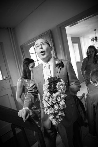 Bruidegom met bloemen kijkt verrast omhoog, gasten op achtergrond bij ontmoeting, zwart-witfoto trouwreportage - Trouwshoot S