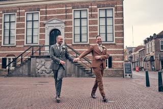Bruidspaar Jeroen en Martijn rennen hand in hand over plein bij historisch bakstenen gebouw, trouwreportage Fletcher Hotel Wi