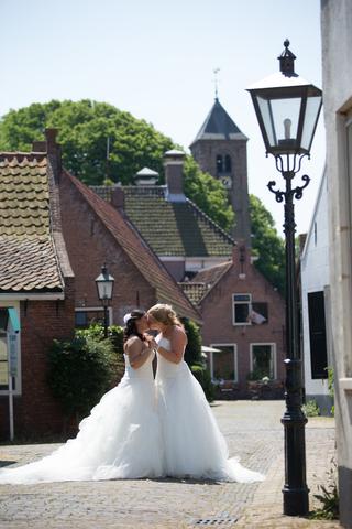 Bruidspaar in witte trouwjurken zoent op plein voor historische kerk, trouwreportage in Nederlands dorpslandschap - Trouwshoo