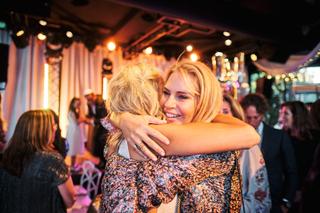 Bruid Liad en gast omhelzen elkaar op trouwfeest The Boathouse Kralingen, festive bruidsfotografie met gouden verlichting - T