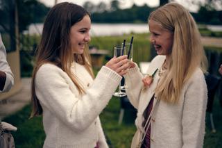 Twee bruidsmeisjes in witte cardigans klinken met champagne in de tuin van Breeze, lachend en vrolijk tijdens de receptie - T