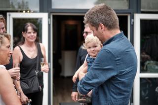 Man houdt blonde peuter vast tijdens jubileumfeest, gasten met glazen op het terras - Trouwshoot Stefan Segers