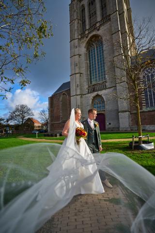 Karlijn en Glenn lopen samen over het kerkplein, haar sluier waaiert mooi uit tegen de imposante kerk van Vierpolders - Trouw