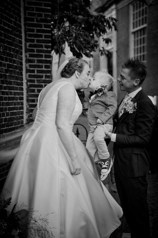 Bruid kust haar kleine zoon terwijl bruidegom toekijkt in de steeg bij Breezze - Trouwshoot Stefan Segers