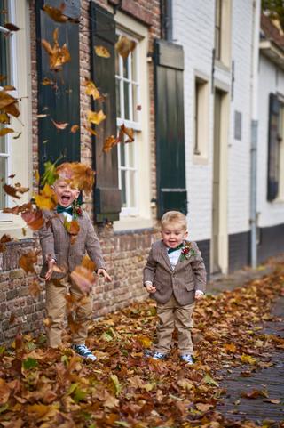Twee bruidsjonkers spelen in herfstbladeren op het terras bij Breezze - pure vreugde tijdens de bruiloft van Karlijn en Glenn