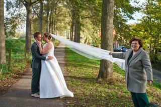 Karlijn en Glenn kussen onder de bomen terwijl een gast lachend de sluier vasthoudt op dit romantische moment in Vierpolders