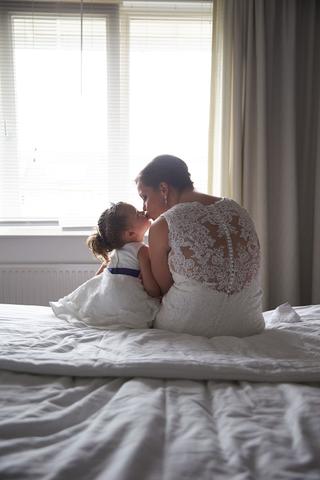 Bruid en bruidsmeisje delen een teder moment op het bed in trouwjurk bij Land van Belofte - Trouwshoot Stefan Segers