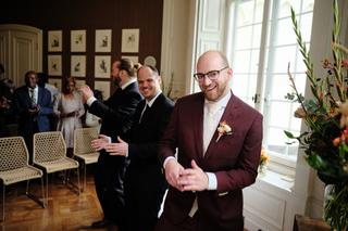 Bruidegom in bordeauxrood pak lacht vol vreugde in de elegant ingerichte zaal van Landgoed De Salentein, gasten op de achterg