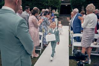 Ringdrager Jayce loopt trots door de ceremonie-aisle met 'Ring Security' bordje terwijl gasten applaudisseren bij Kasteel De