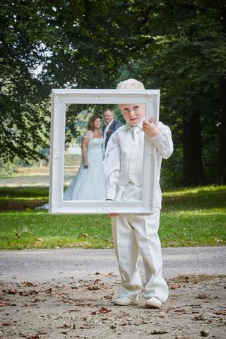 Ringjongen Joshua in wit pak houdt decoratief frame omhoog met bruidspaar Eric en Jessica achter zich - Trouwshoot Stefan Seg