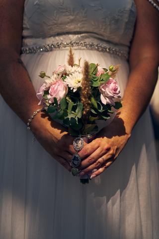 Bruidsbouquet met roze rozen en witte dahlia's in handen van Diliana, met herinnerings-medaillons en trouwring zichtbaar in z