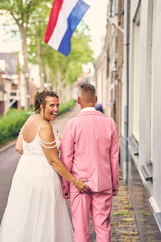 Diliana en Olav wandelen hand in hand door een Nederlandse straat onder de Nederlandse vlag, bruid in wit met pareldetails ki