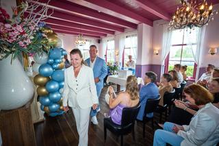 Daphne in witte blazer loopt door de feestzaal onder blauwe en gouden ballonnen terwijl gasten applaudisseren op Hoeve Kromwi